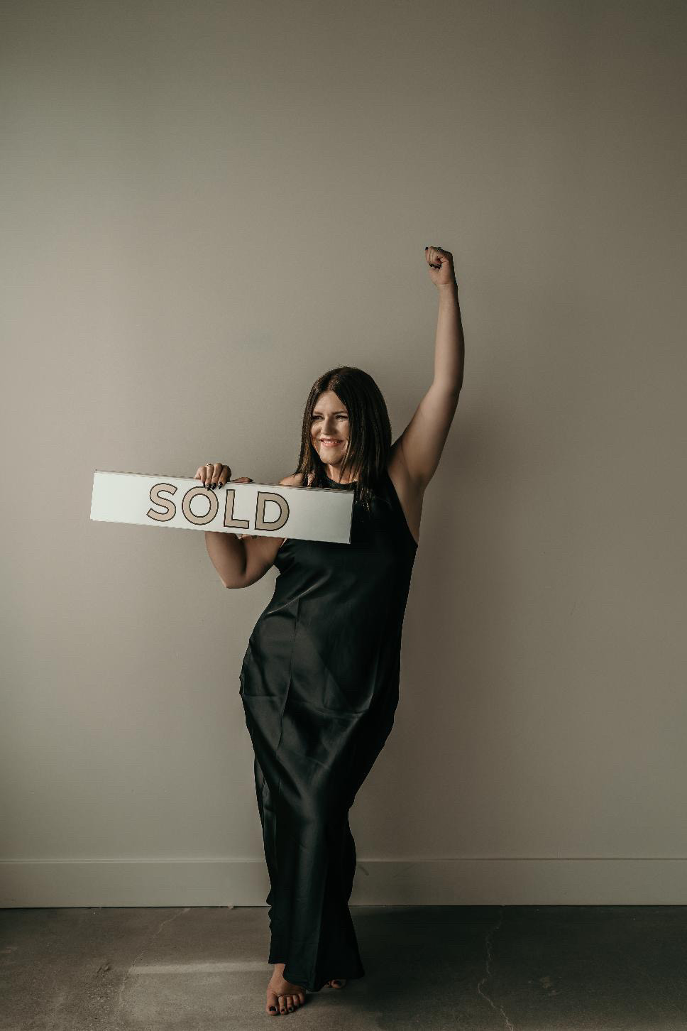 Woman in a black dress holding a "SOLD" sign with one arm raised in celebration.