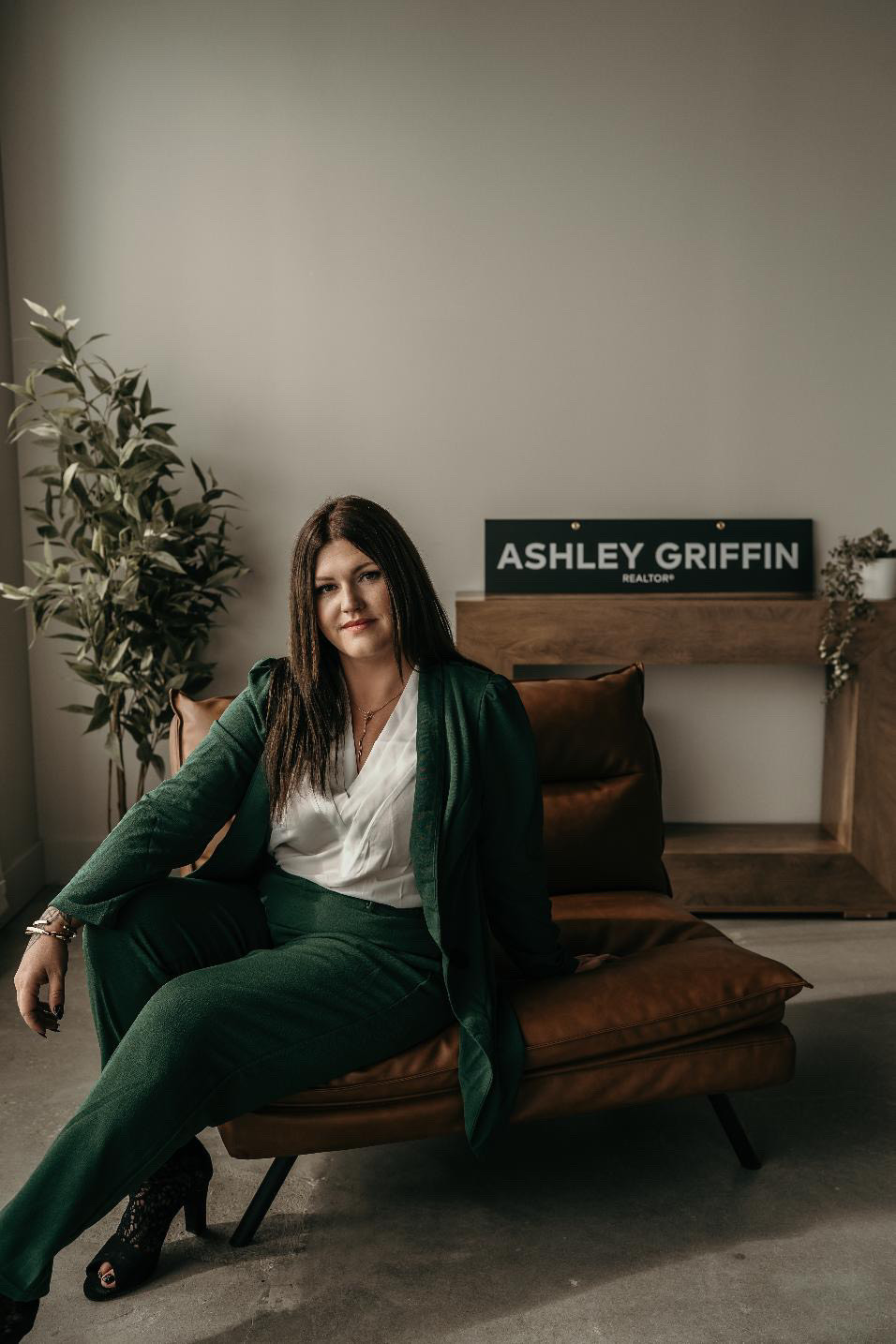 Professional real estate agent in green suit sitting confidently on a leather chair with a name sign reading "Ashley Griffin Realtor."