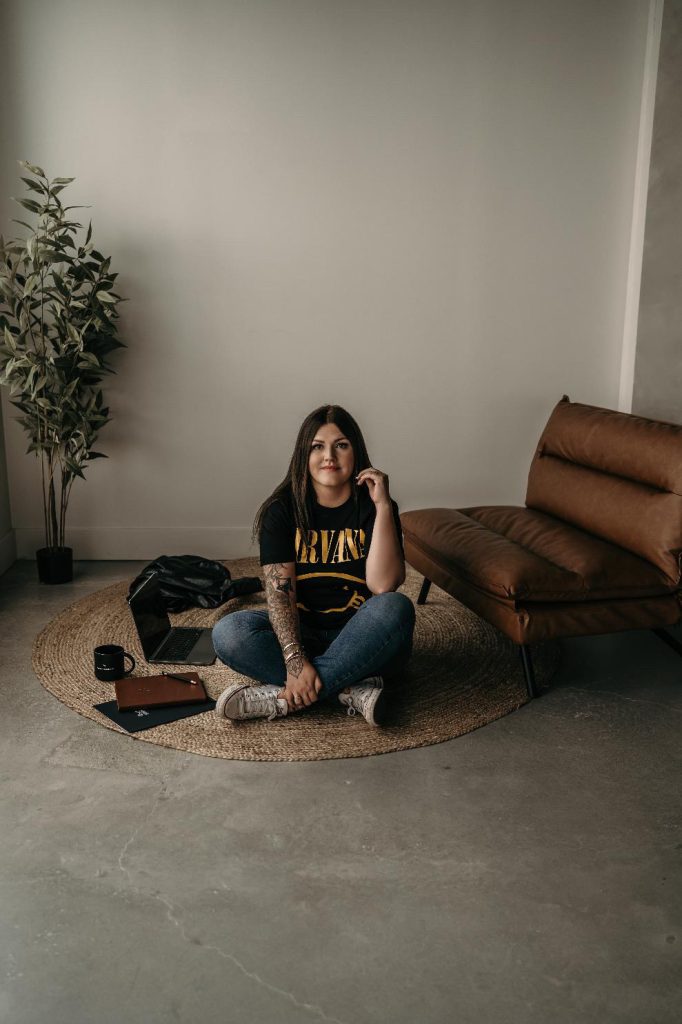 Casual real estate agent sitting on round rug in a Nirvana t-shirt with laptop, notebook and coffee mug nearby.